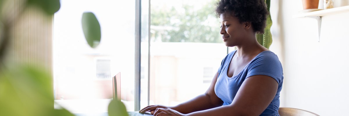 A person sitting at a desk in a sunny home office and typing on a laptop.