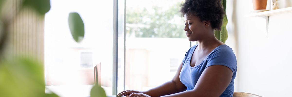 A person sitting at a desk in a sunny home office and typing on a laptop.
