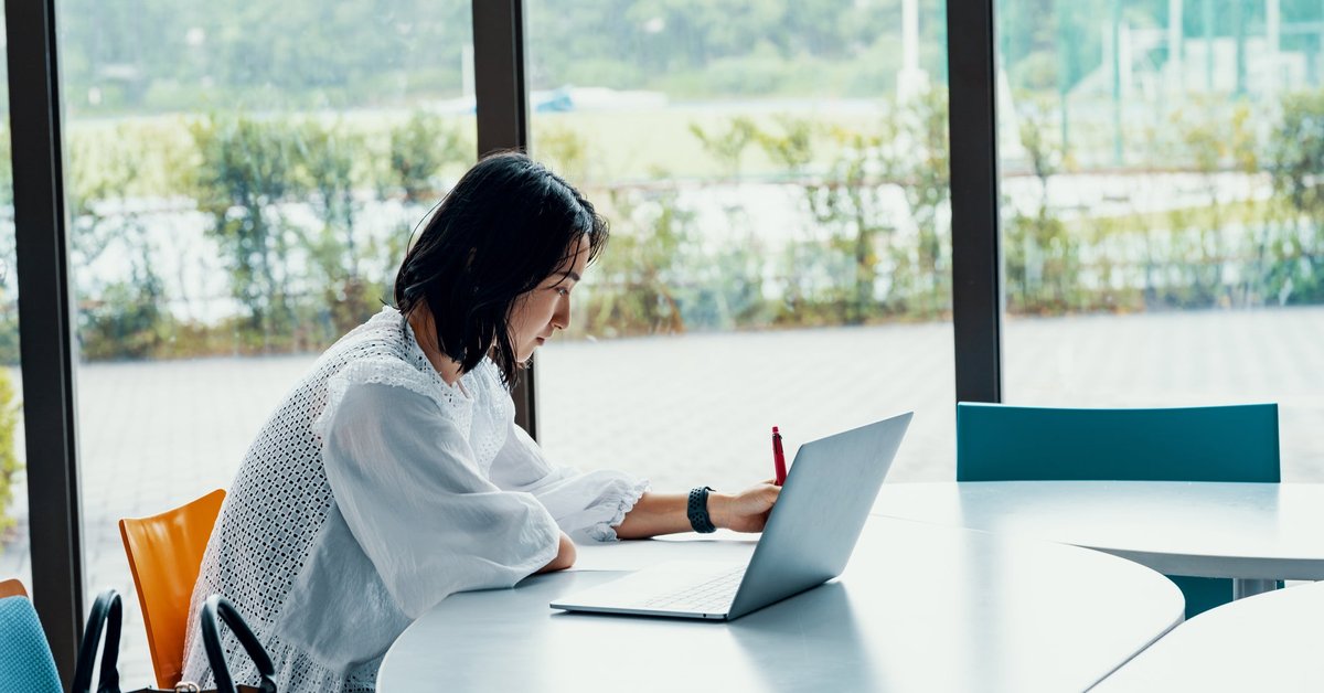 A person sitting at a table next to a big window and writing in a notebook next to an open laptop.