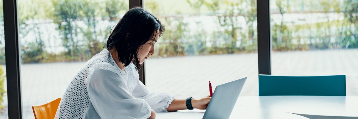 A person sitting at a table next to a big window and writing in a notebook next to an open laptop.