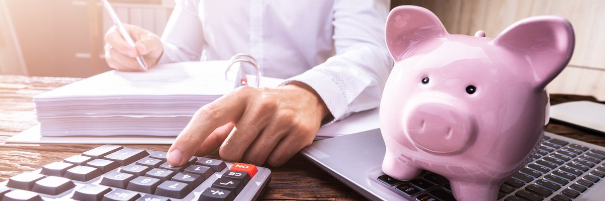 Person makes calculations and writes them in a notebooks while a piggy bank sits nearby.
