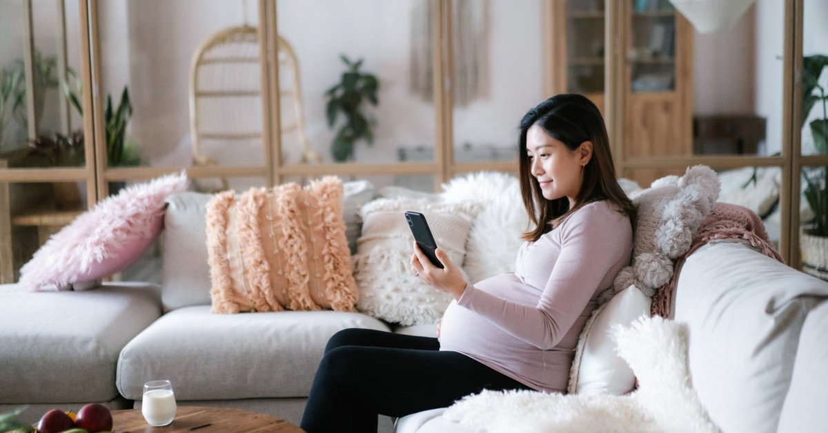 A pregnant person sitting on their couch and looking at their phone.