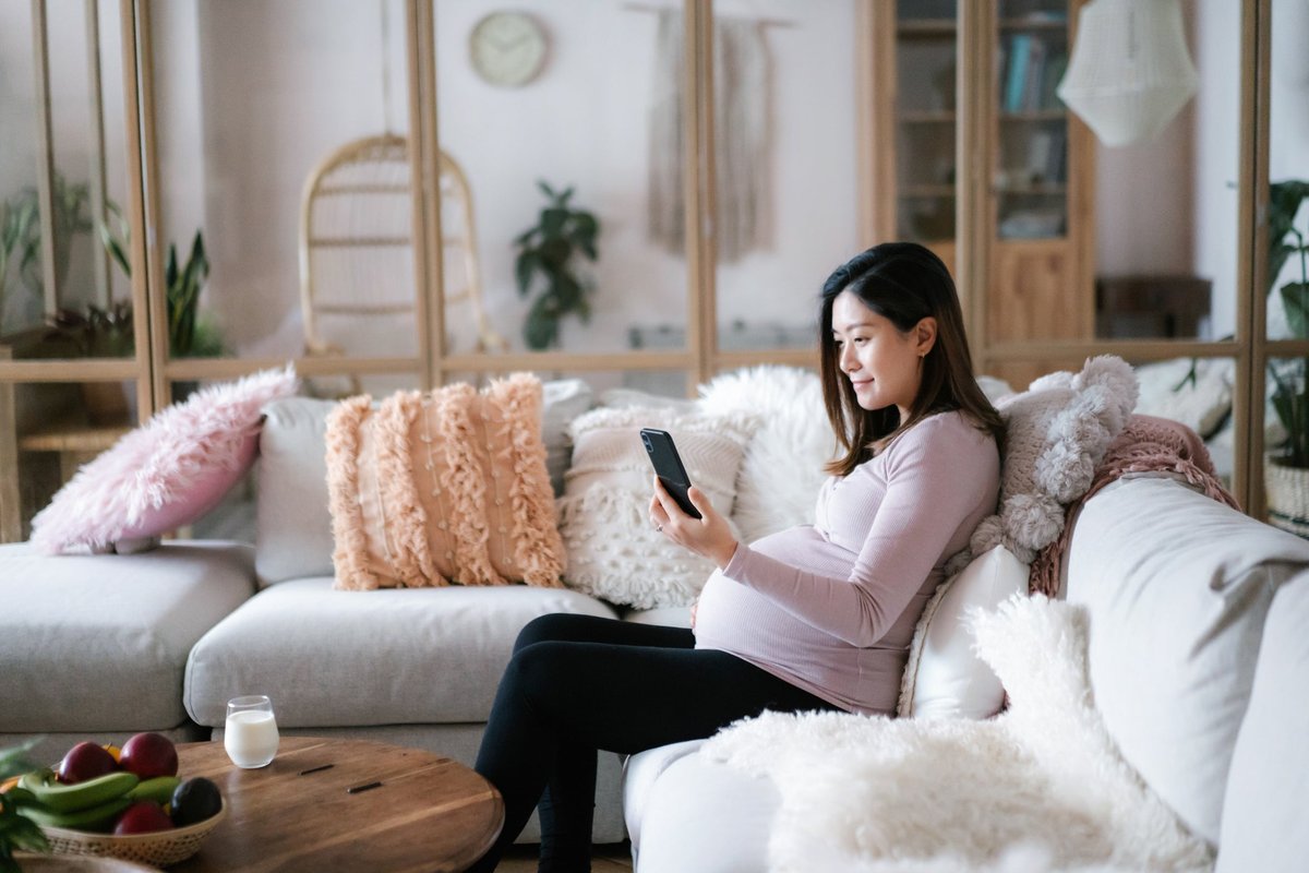 A pregnant person sitting on their couch and looking at their phone.
