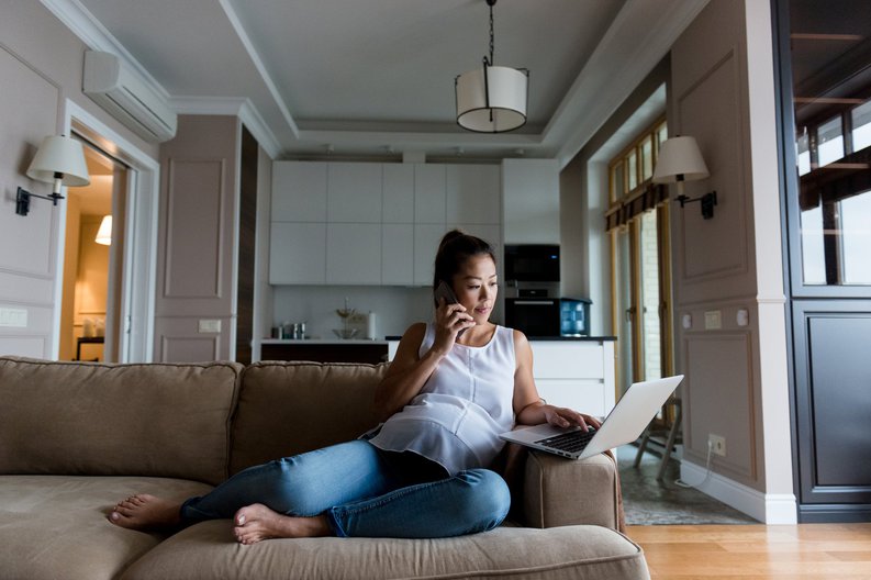 A pregnant woman sitting on her couch while searching on her computer and making a phone call.