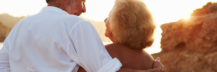 An older couple standing on the beach and looking at the sunset with their arms around each other.