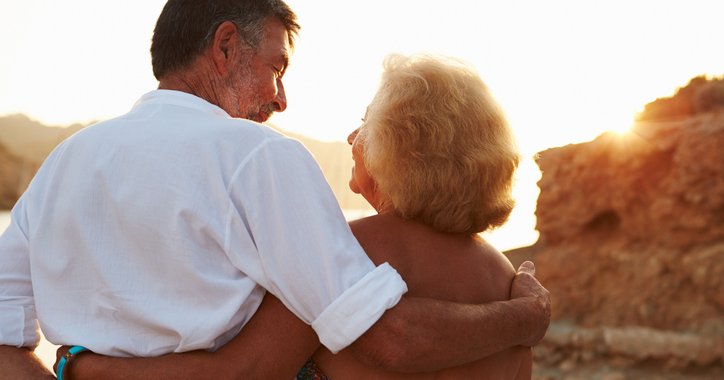 An older couple standing on the beach and looking at the sunset with their arms around each other.