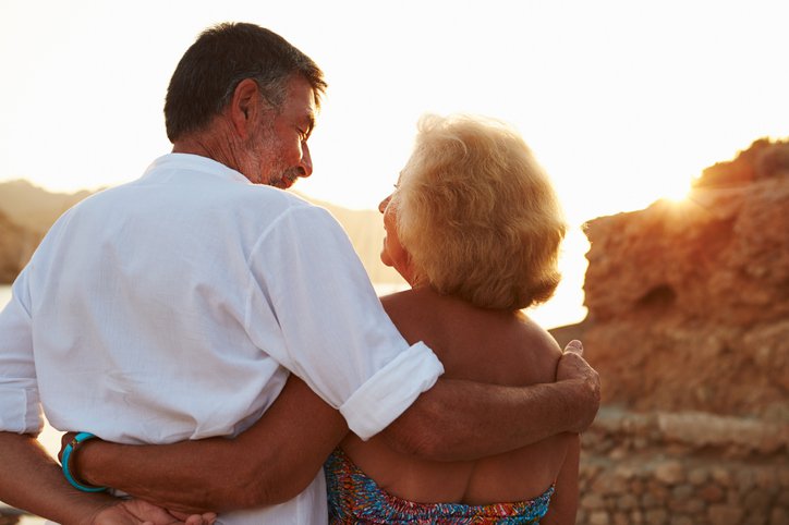 An older couple standing on the beach and looking at the sunset with their arms around each other.