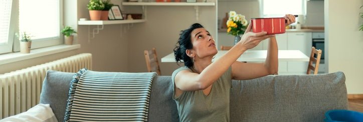 A woman sitting on the couch catches a water leak with a cooking pot.