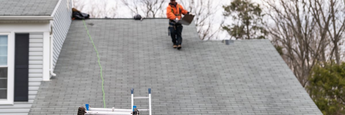 A roofer walking on top of a house and a truck with ladders parked out front.