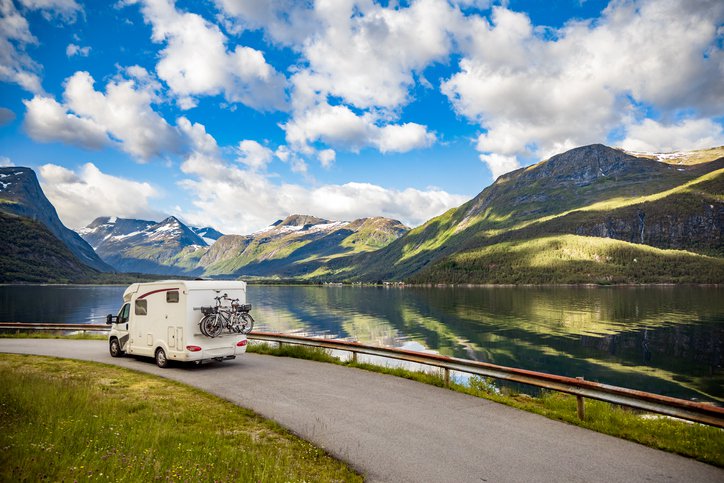 An RV driving along a road beside a mountain lake reflecting the blue sky and clouds.