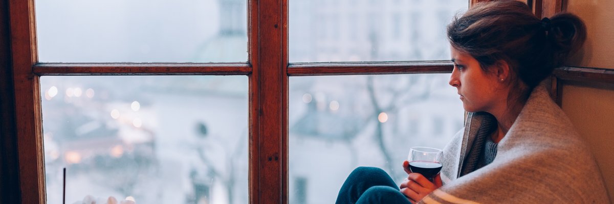 A worried woman wrapped in a blanket sitting at a window.