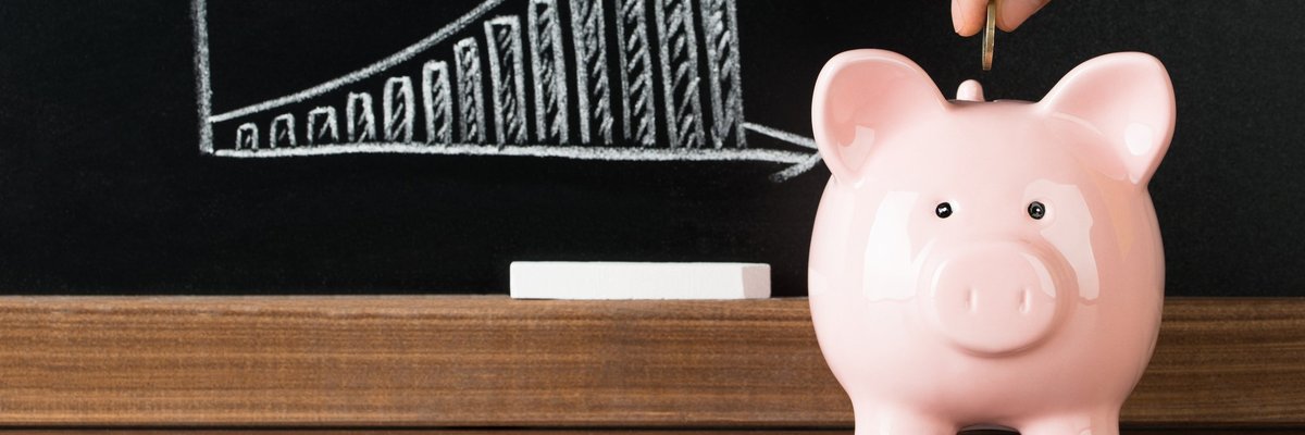 Coin being inserted into pink piggy bank in front of blackboard with rising graph