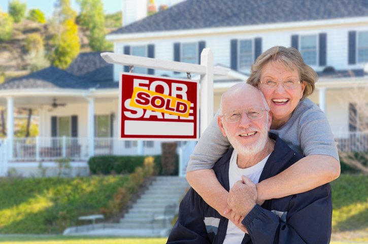 A senior couple in front of a house and a "sold" sign.