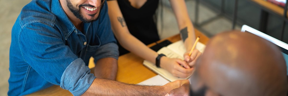 Two people meeting with a third person in an office and shaking hands across the desk.