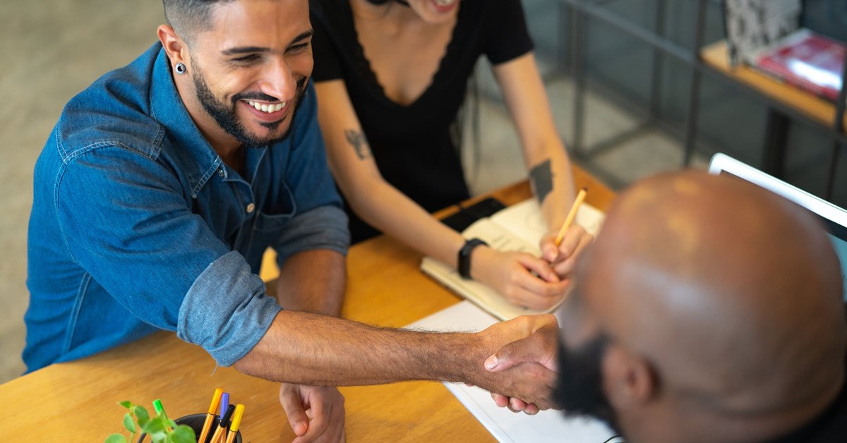 Two people meeting with a third person in an office and shaking hands across the desk.