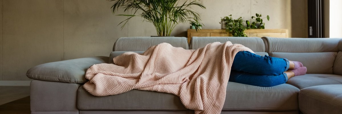 A sick person laying on their couch at home covered by a blanket.
