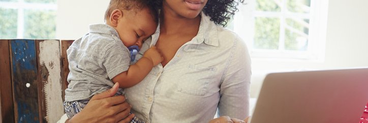 A mother holds an infant while she works on her computer.