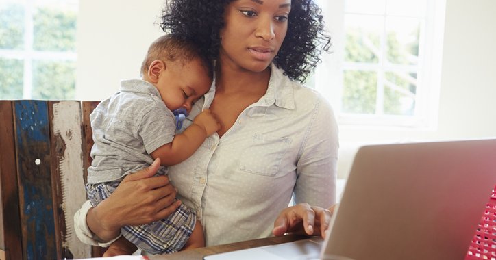 A mother holds an infant while she works on her computer.