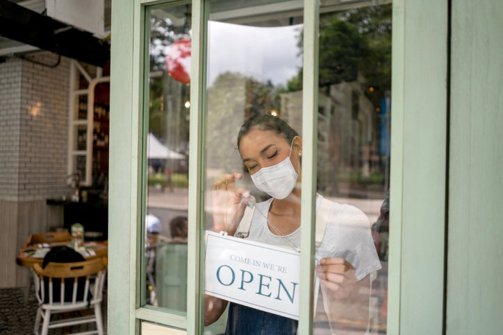 A small business owner wears a mask and hangs an Open sign on the front door of his store.