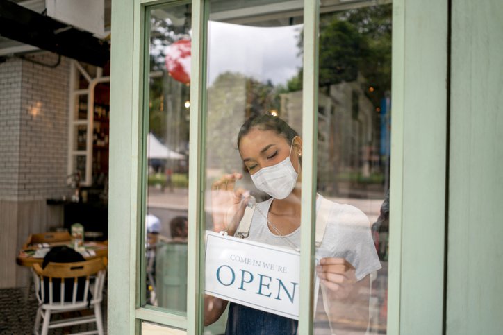 A small business owner wearing a mask and hanging an Open sign on the front door of her shop.