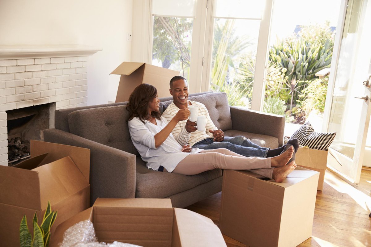 smiling couple toasting with coffee mugs on couch in empty apartment amid moving boxes
