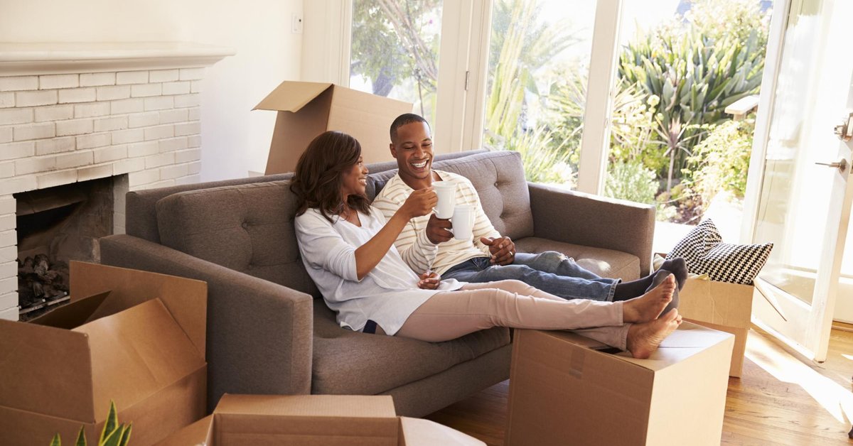 smiling couple toasting with coffee mugs on couch in empty apartment amid moving boxes