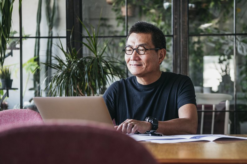 A smiling man working on a laptop in a room full of plants.
