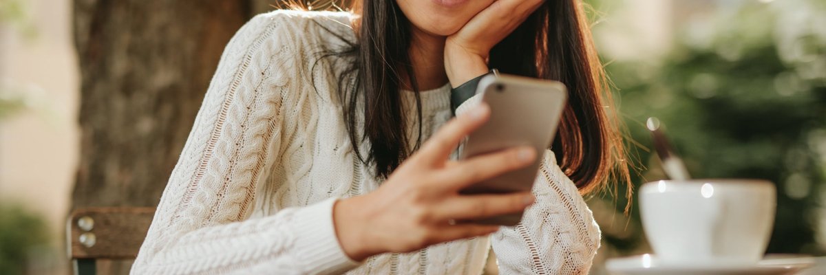 A smiling person sitting at an outdoor cafe table and looking at their cellphone.