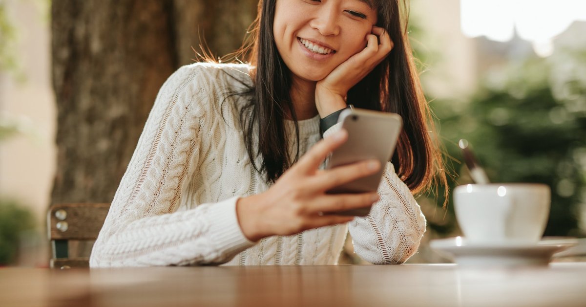 A smiling person sitting at an outdoor cafe table and looking at their cellphone.