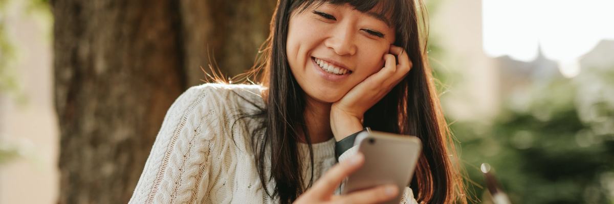 A smiling person sitting at an outdoor cafe table and looking at their cellphone.