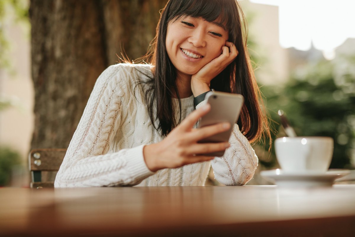 Une personne souriante assise à une table de café en plein air et regardant son téléphone portable.