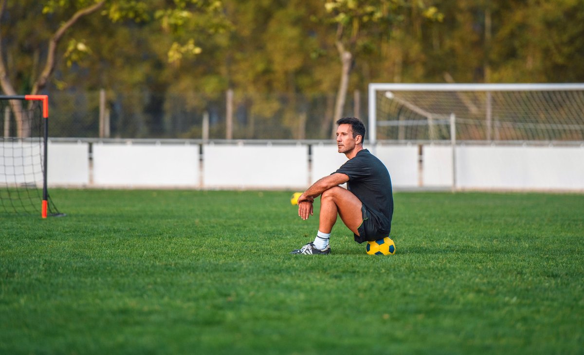 A soccer player sits on a ball in the middle of the field, looking pensive.