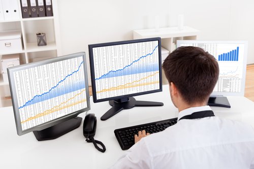A man using a computer with three different monitors full of charts and graphs.