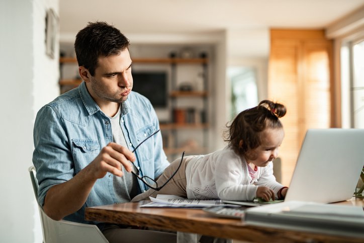 A stressed man trying to work from his kitchen table at home while his baby crawls across his laptop.