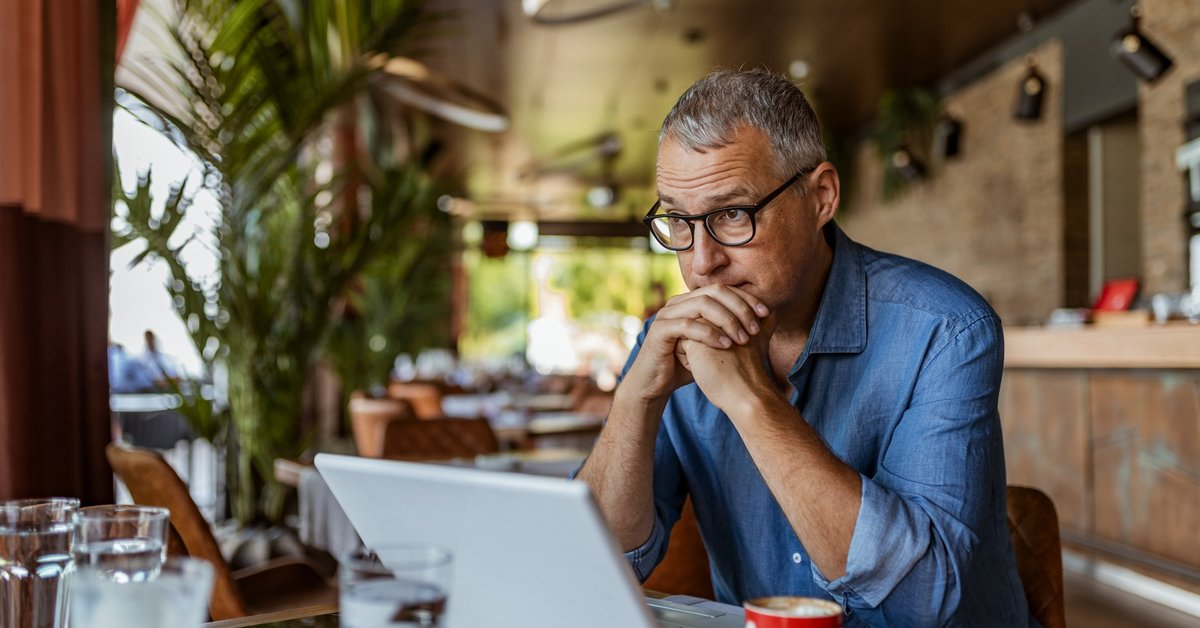 A man sitting in front of his laptop at a cafe table and looking stressed.