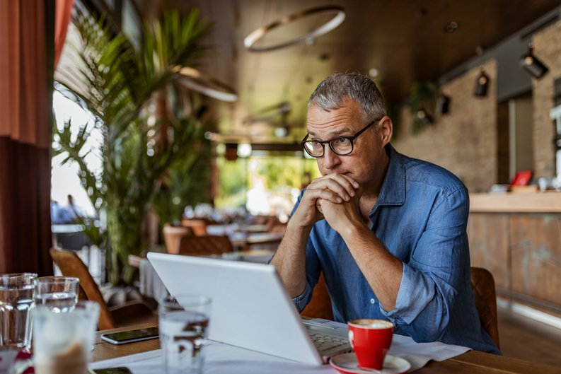 A man sitting in front of his laptop at a cafe table and looking stressed.