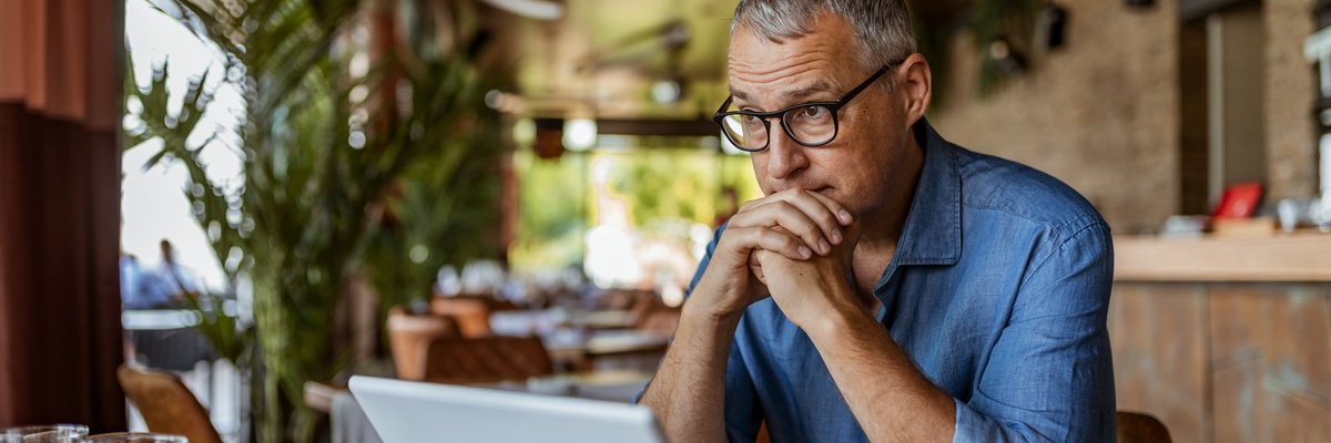A man sitting in front of his laptop at a cafe table and looking stressed.
