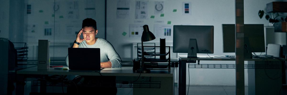 A stressed young man sitting in front of a laptop in a dark office.
