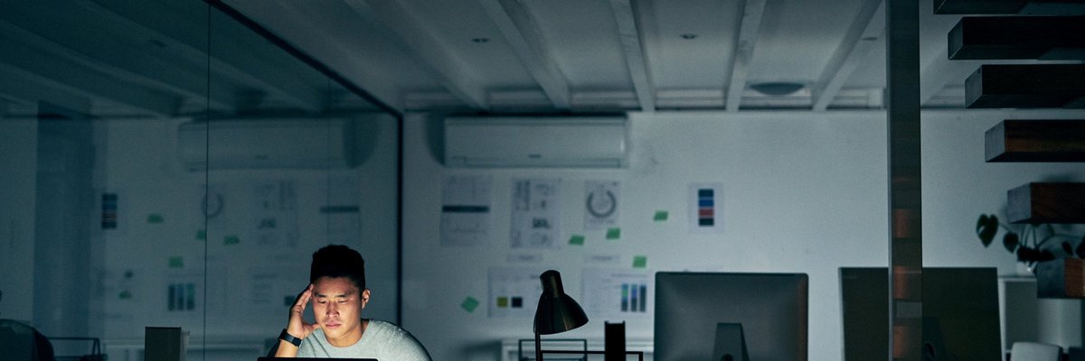 A stressed-looking man staring at his computer while sitting alone in a dark office.