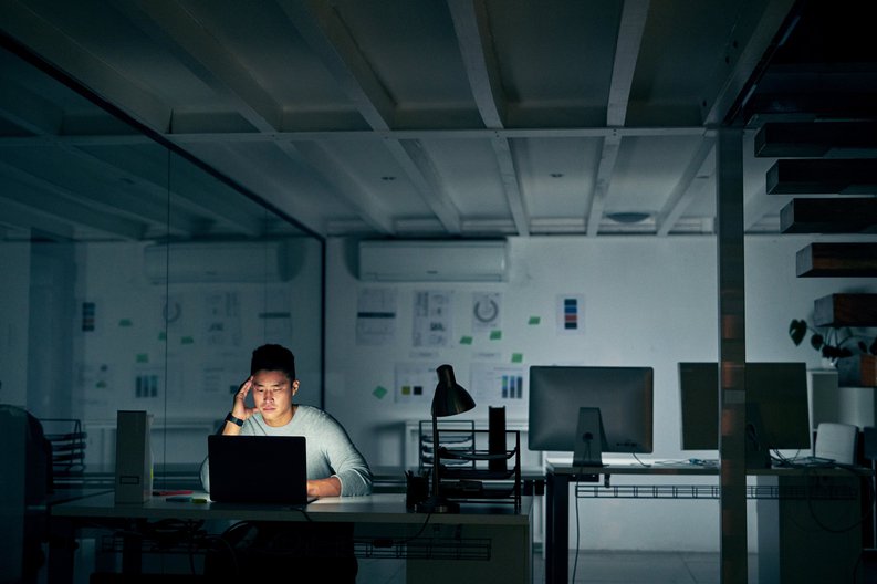 A stressed young man sitting in front of a laptop in a dark office.