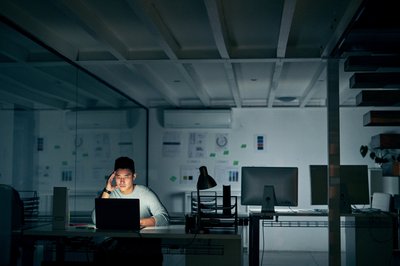 A stressed-looking man staring at his computer while sitting alone in a dark office.