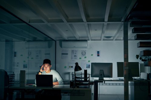 A stressed-looking man staring at his computer while sitting alone in a dark office.