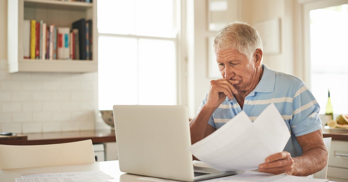 A stressed person looking through papers and sitting in front of a laptop at a kitchen counter.
