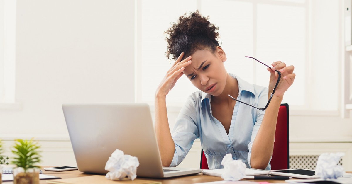 Woman at laptop holding her head; on the table in front of her are crumbled up pieces of paper