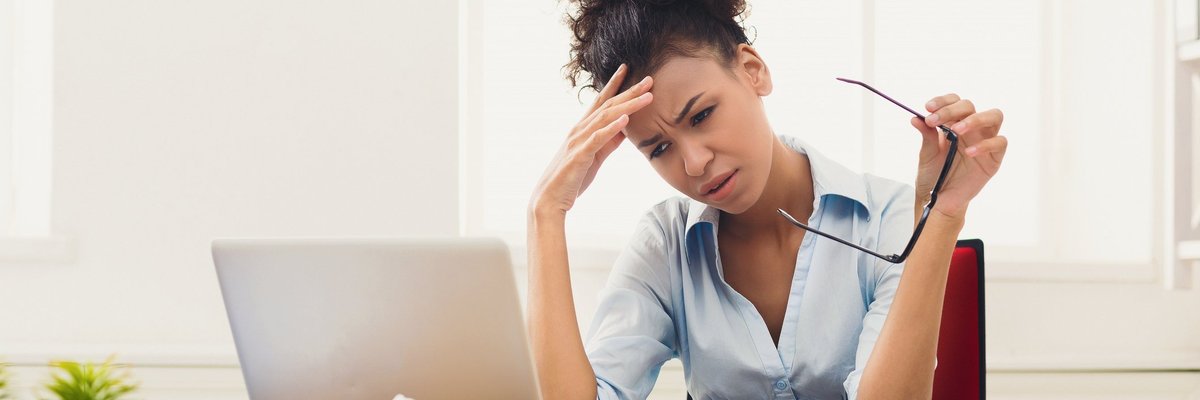 Woman at laptop holding her head; on the table in front of her are crumbled up pieces of paper