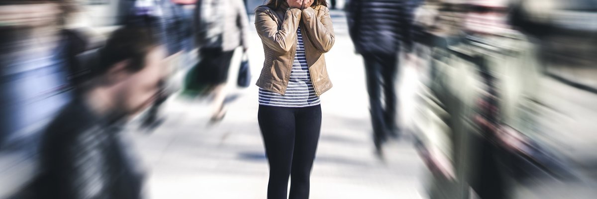 A stressed young woman in a busy crowd covering her face with her hands.