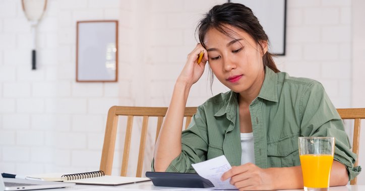 A stressed woman sitting at her dining table and looking through her bills and receipts with her head resting in her hand.