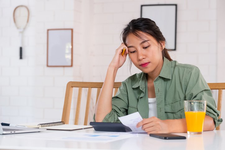 A stressed woman sitting at her dining table and looking through her bills and receipts with her head resting in her hand.