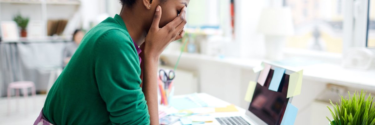 A stressed young woman sitting at her desk in front of a laptop with her face in her hands.