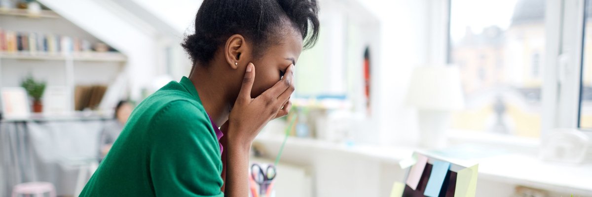 A stressed young woman sitting at her desk in front of a laptop with her face in her hands.
